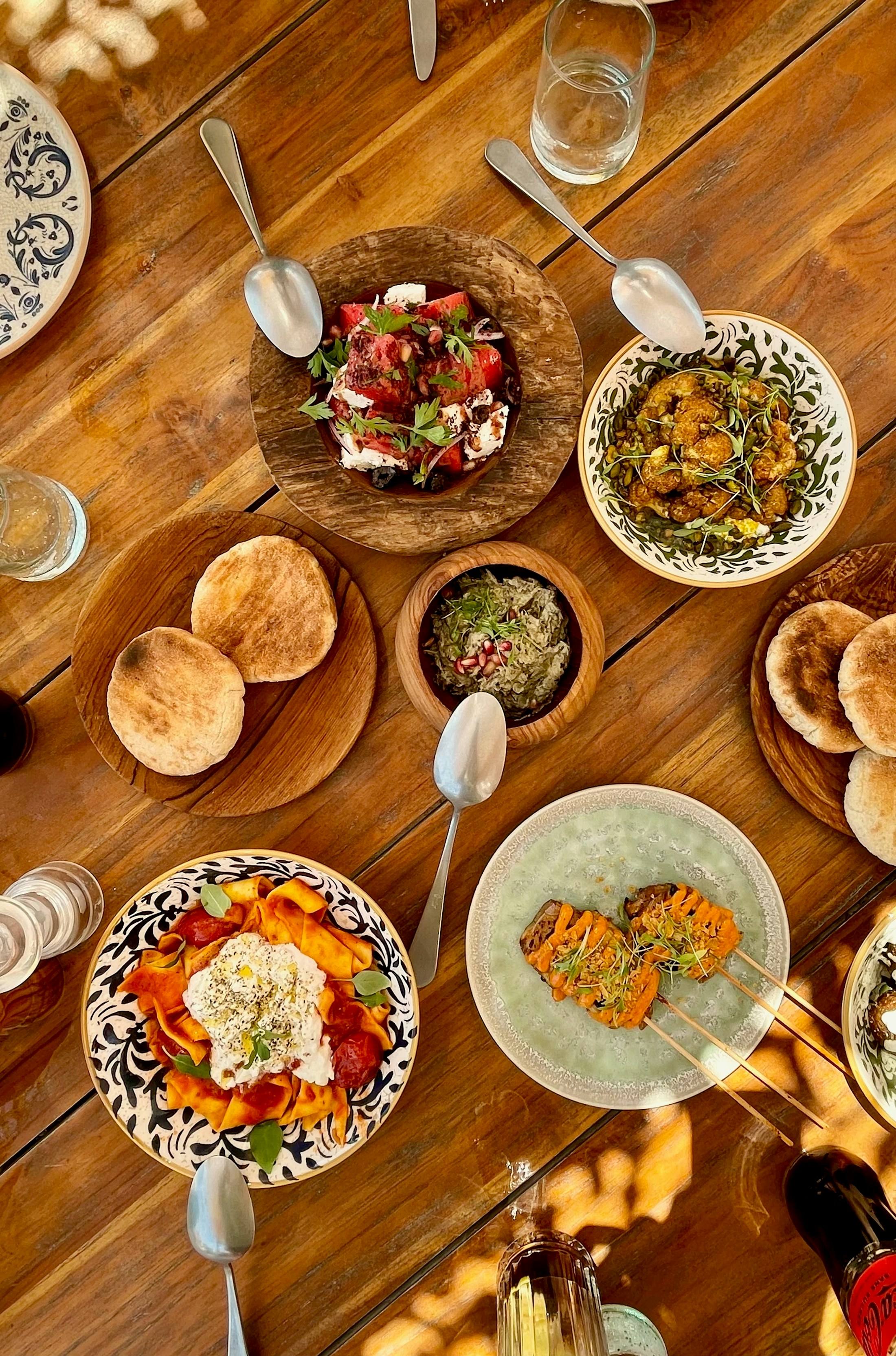 Een houten tafel vol gedeelde gerechten, brood en salades, van bovenaf gezien.